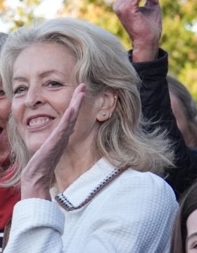 A woman with blonde hair in a cream jacket, hand raised, smiling amid the crowd at the Piddington announcement